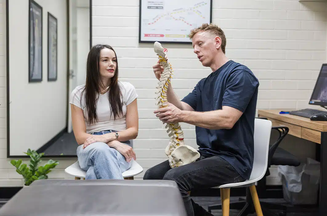 Man explaining spine model to woman seated in medical consultation room with computer and charts.