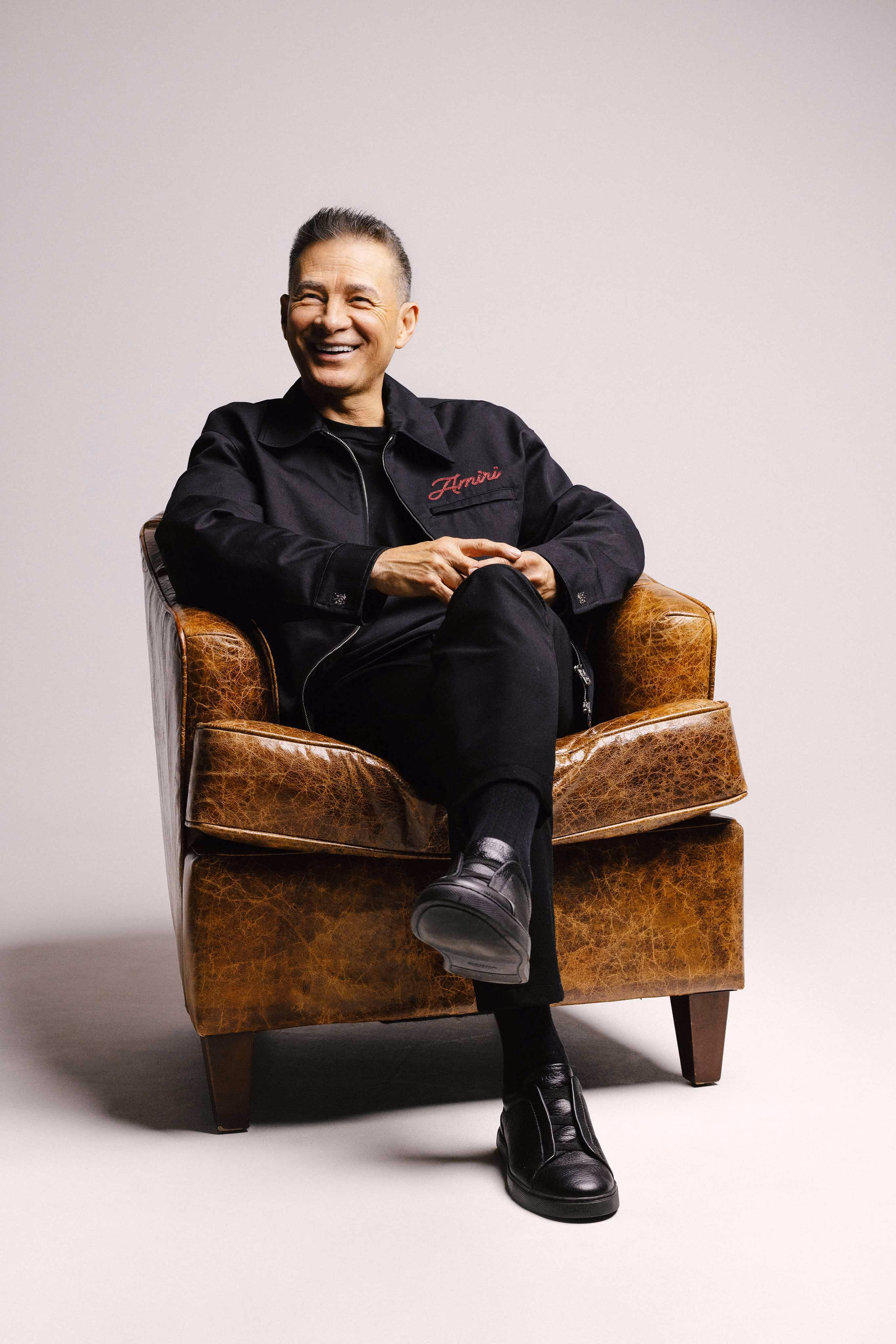 Hombre sonriente sentado con chaqueta negra y zapatos negros en un sillón marrón de cuero.
