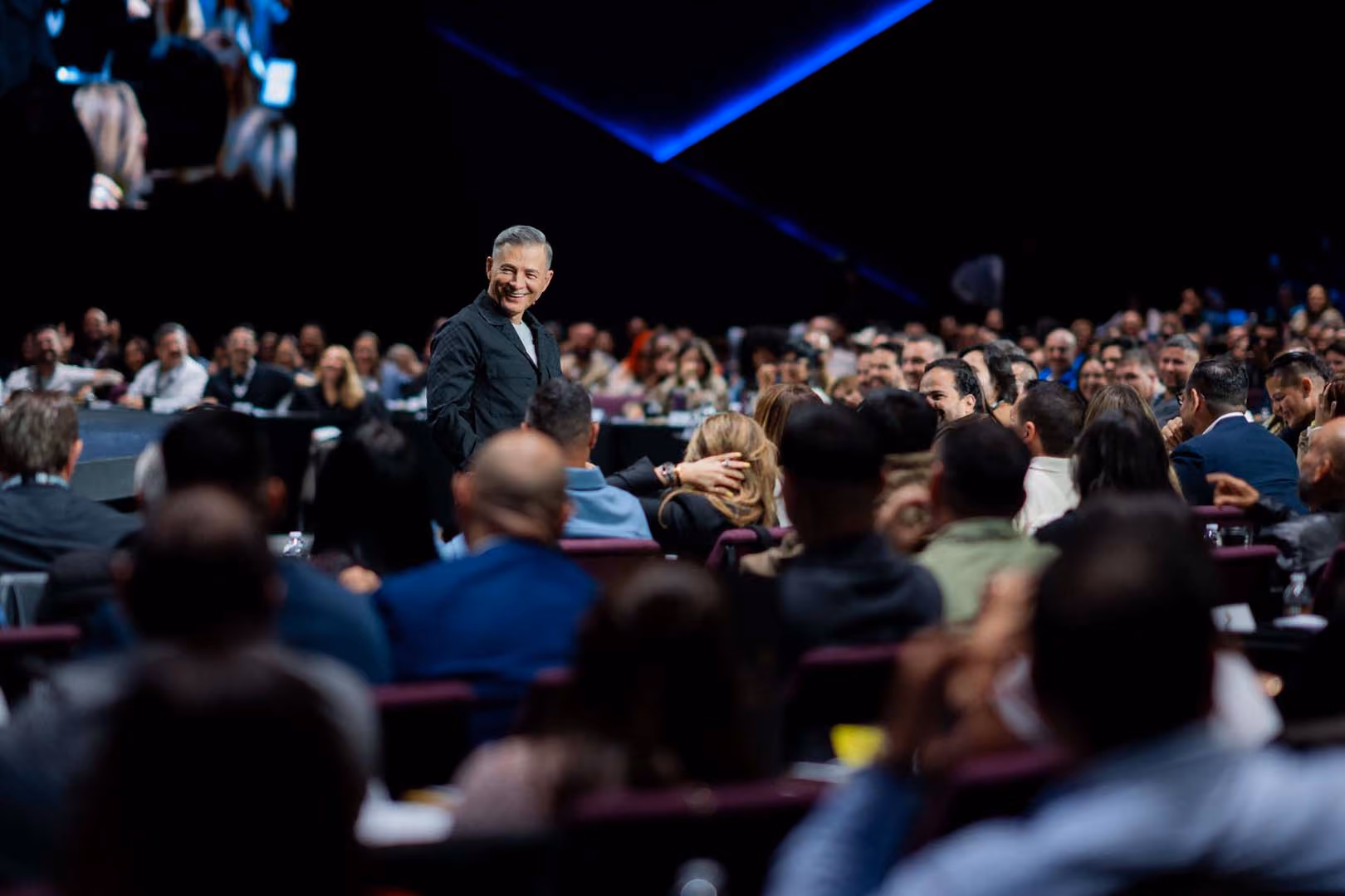 Un hombre con saco negro sonriente está de pie en medio de una gran audiencia sentada en un auditorio oscuro con luces azules, mientras la multitud presta atención.