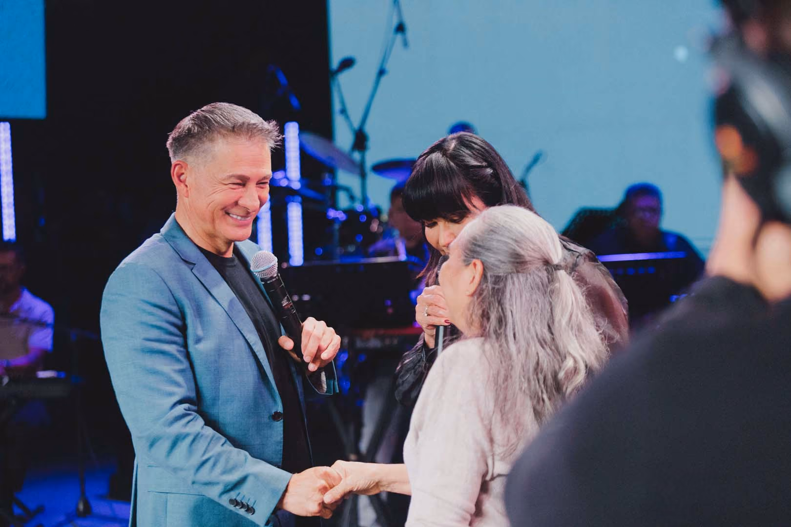 Hombre sonriente con saco azul sostiene micrófono y da la mano a mujer de cabello canoso mientras otra mujer habla con micrófono en un escenario con luces y músicos al fondo.