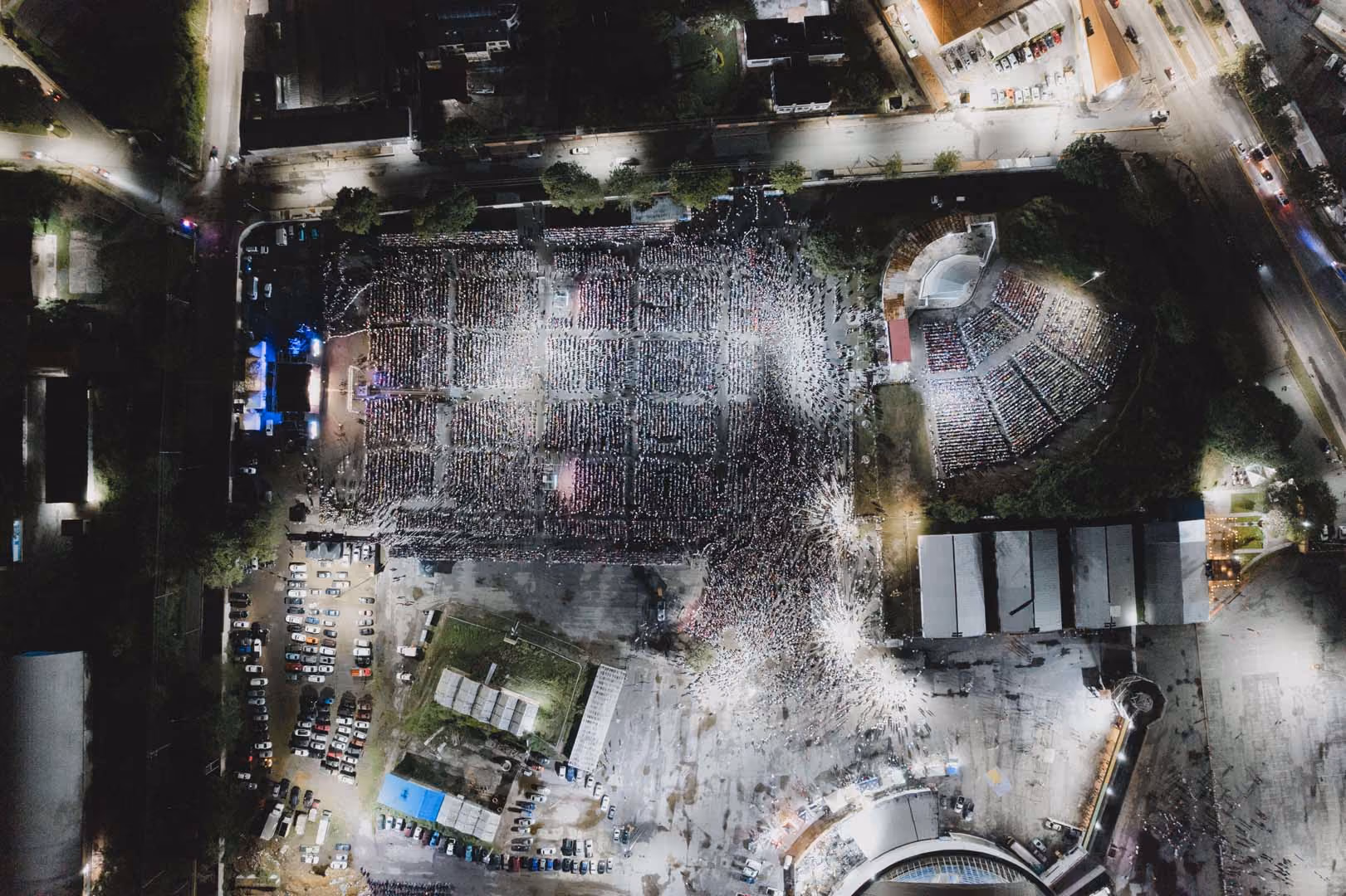 Vista aérea nocturna de un gran estadio con miles de personas reunidas en el campo y las gradas, iluminadas por luces brillantes.