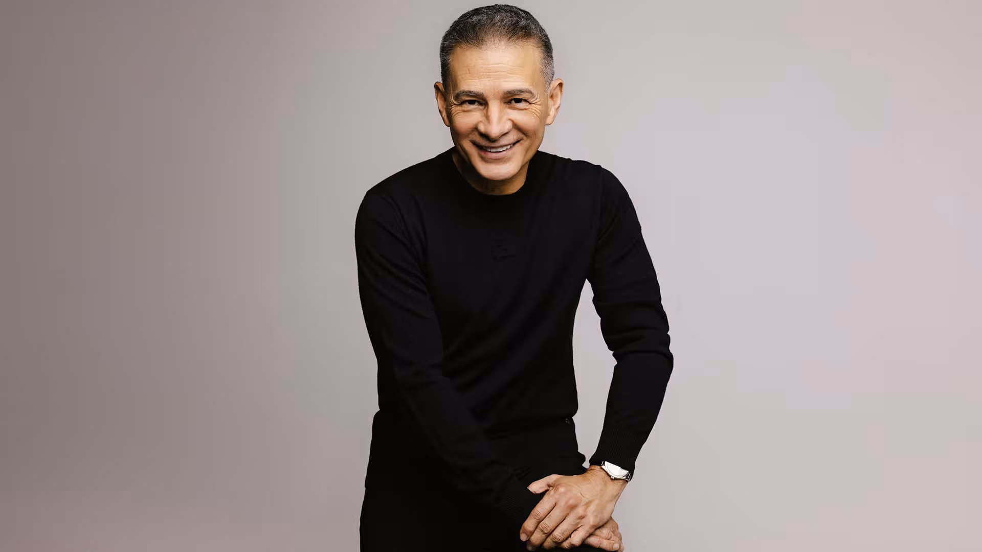 Hombre de mediana edad sonriente con suéter negro y reloj de pulsera, posando sobre fondo neutro claro.