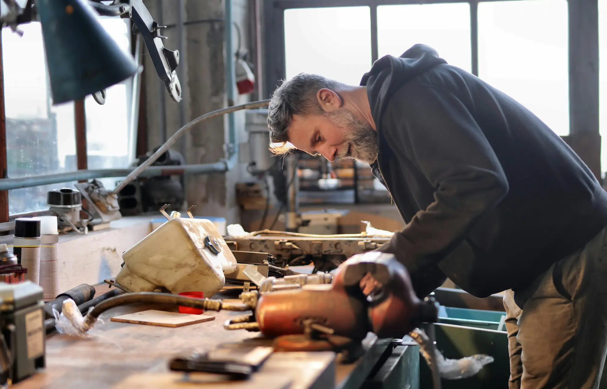 Man with gray beard working on parts at a cluttered workbench in a workshop with large windows.