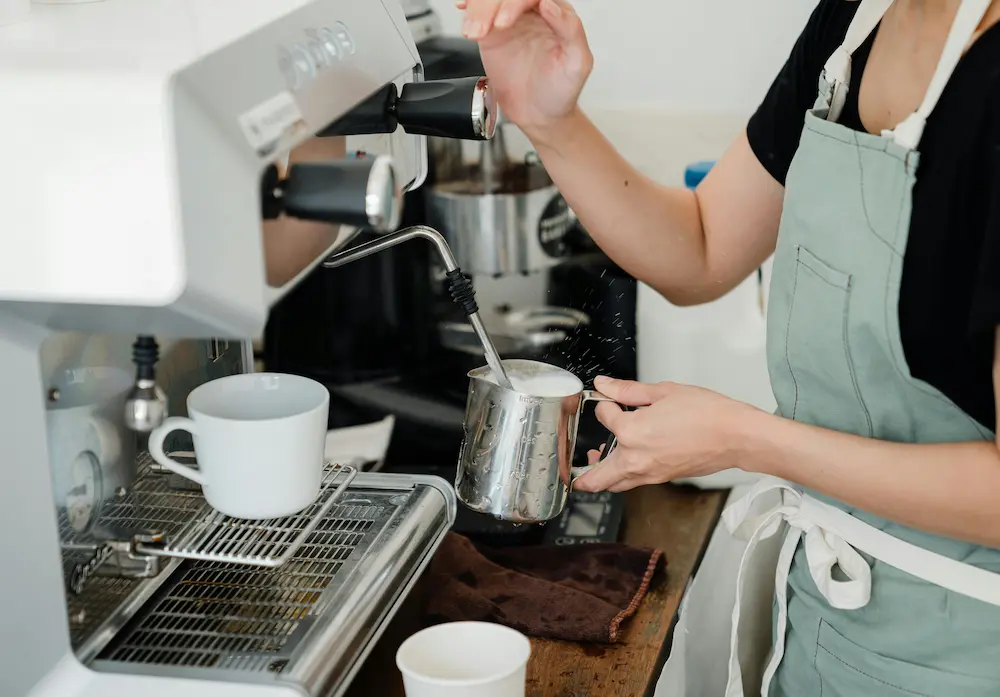 Barista frothing milk with a steam wand next to an espresso machine with a white cup.