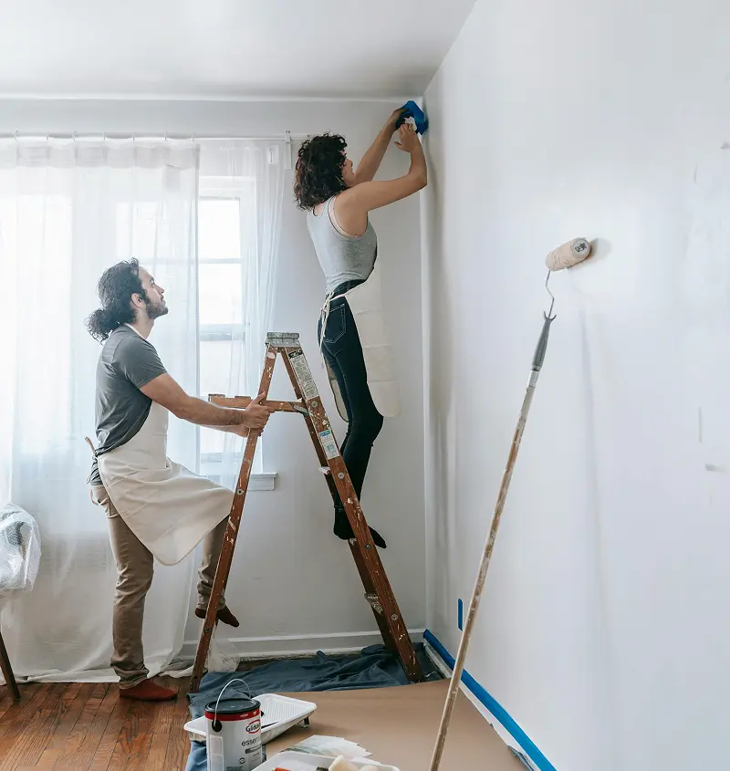 Couple painting a room: woman on ladder applying tape, man steadying ladder, with painting tools below.