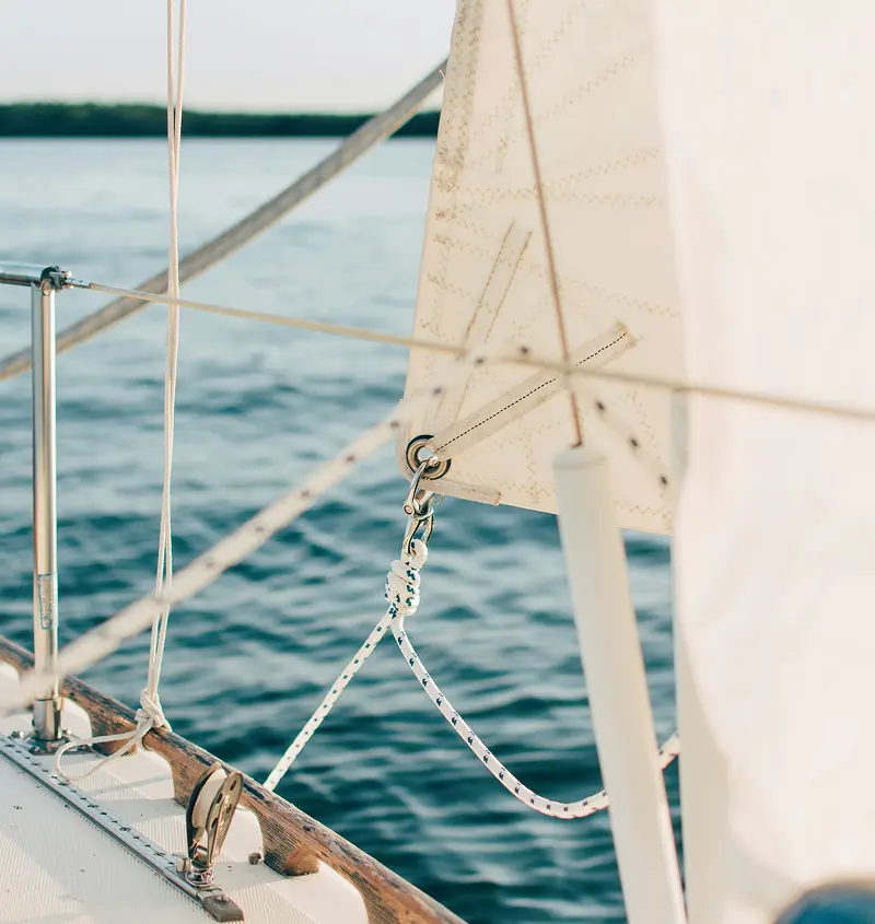 Close-up of sailboat rigging and white sail with calm water and distant shore in the background.