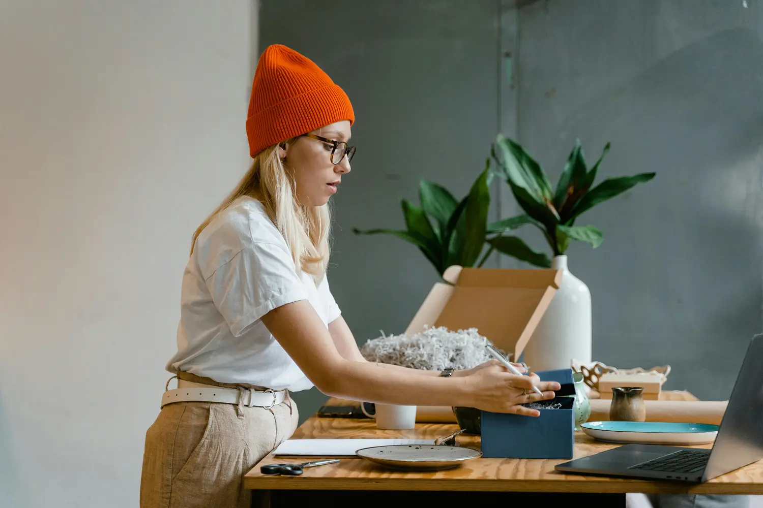 Woman wearing an orange beanie and glasses opening a blue box on a wooden table with laptop and plants.