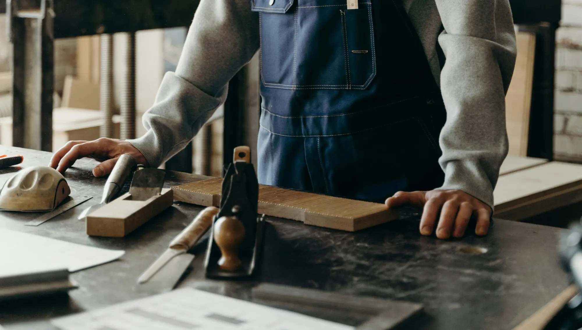Person wearing blue apron standing at a workshop bench with woodworking tools and a wooden plank.