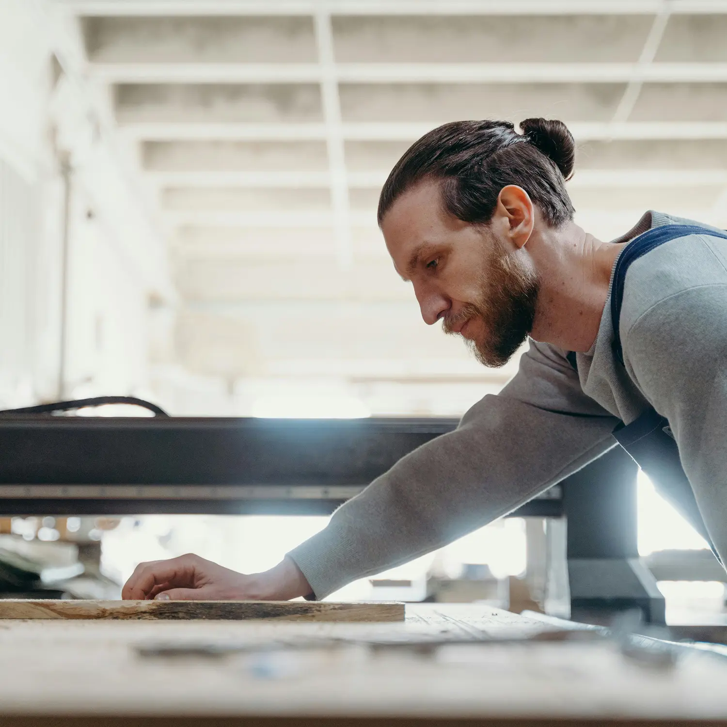 Man with a bun inspecting a piece of wood on a workbench in a bright workshop.