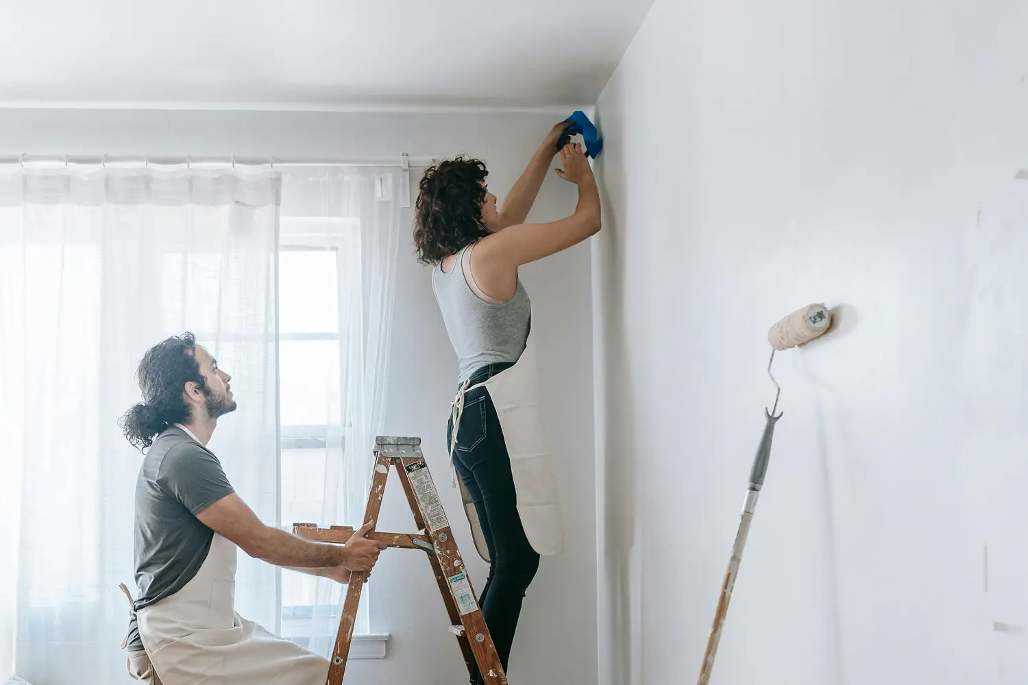 Woman standing on ladder taping a wall corner while man holds the ladder, preparing to paint a room.