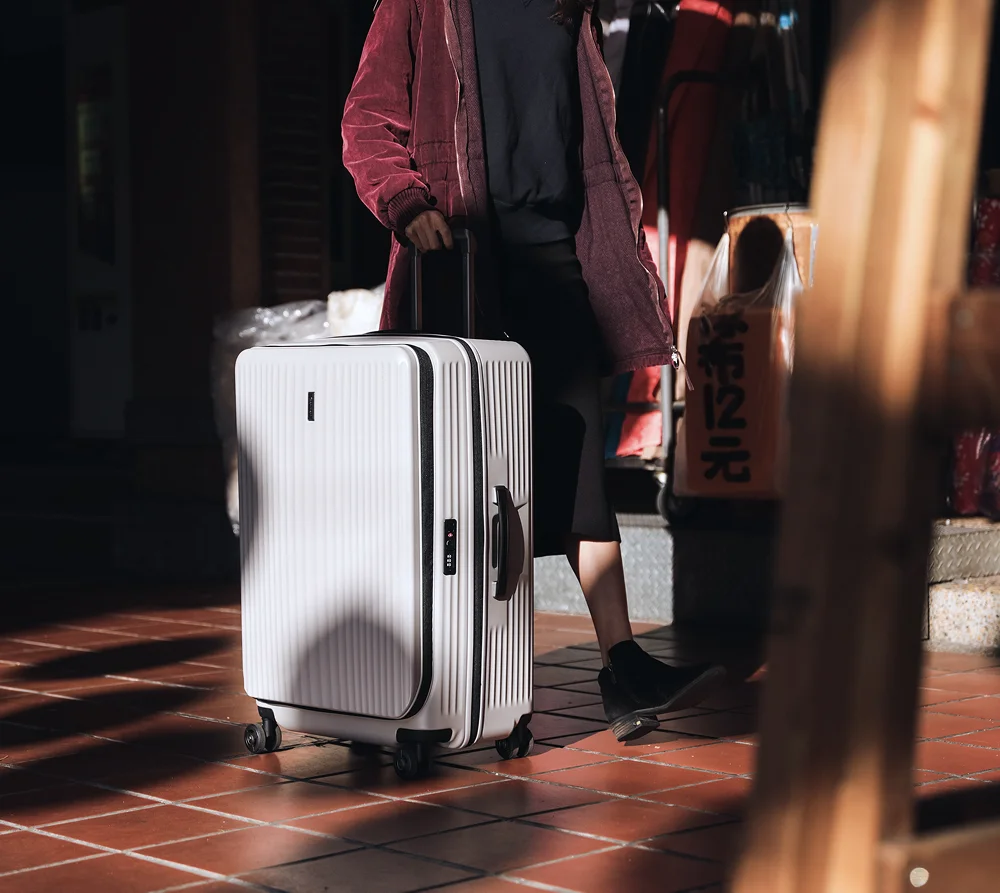 Person in burgundy jacket pulling a large white hard-shell suitcase with wheels on tiled floor.