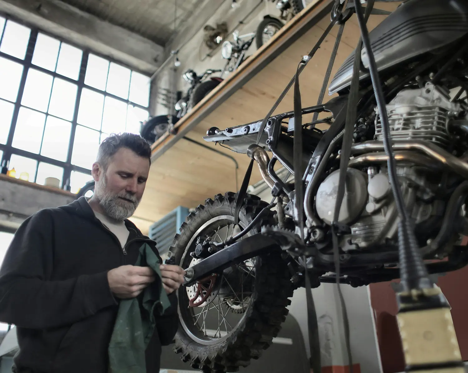 Man with beard cleaning or repairing a suspended dirt bike in a workshop with large windows.