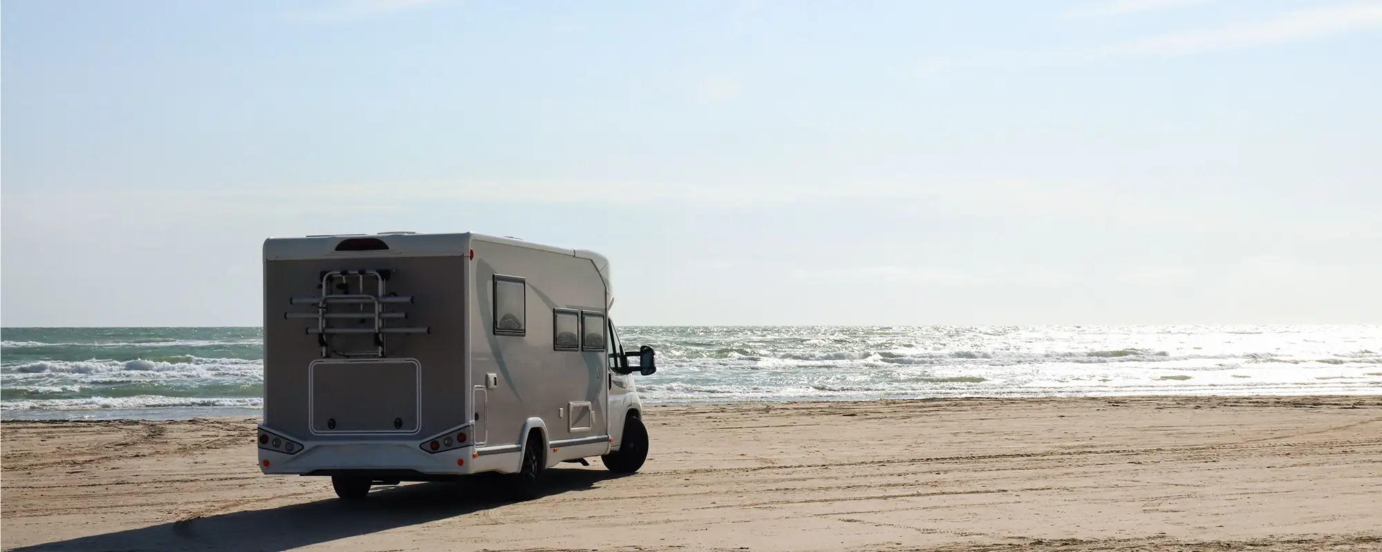 White caravan parked on a sandy beach with ocean waves and clear sky in the background.