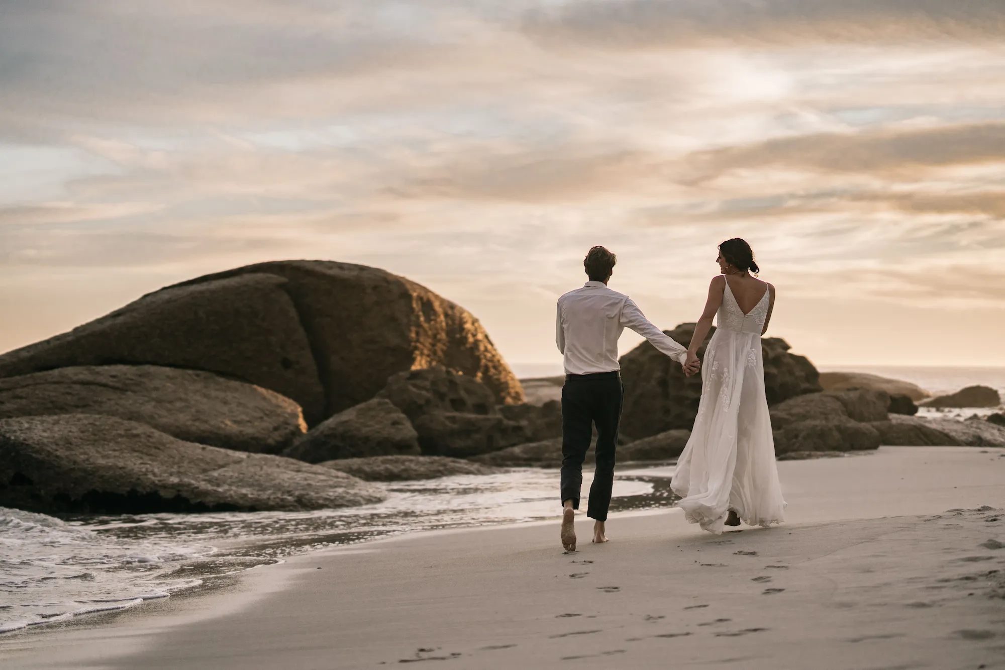 A couple in wedding attire holding hands walking barefoot on a sandy beach near large rocks at sunset.