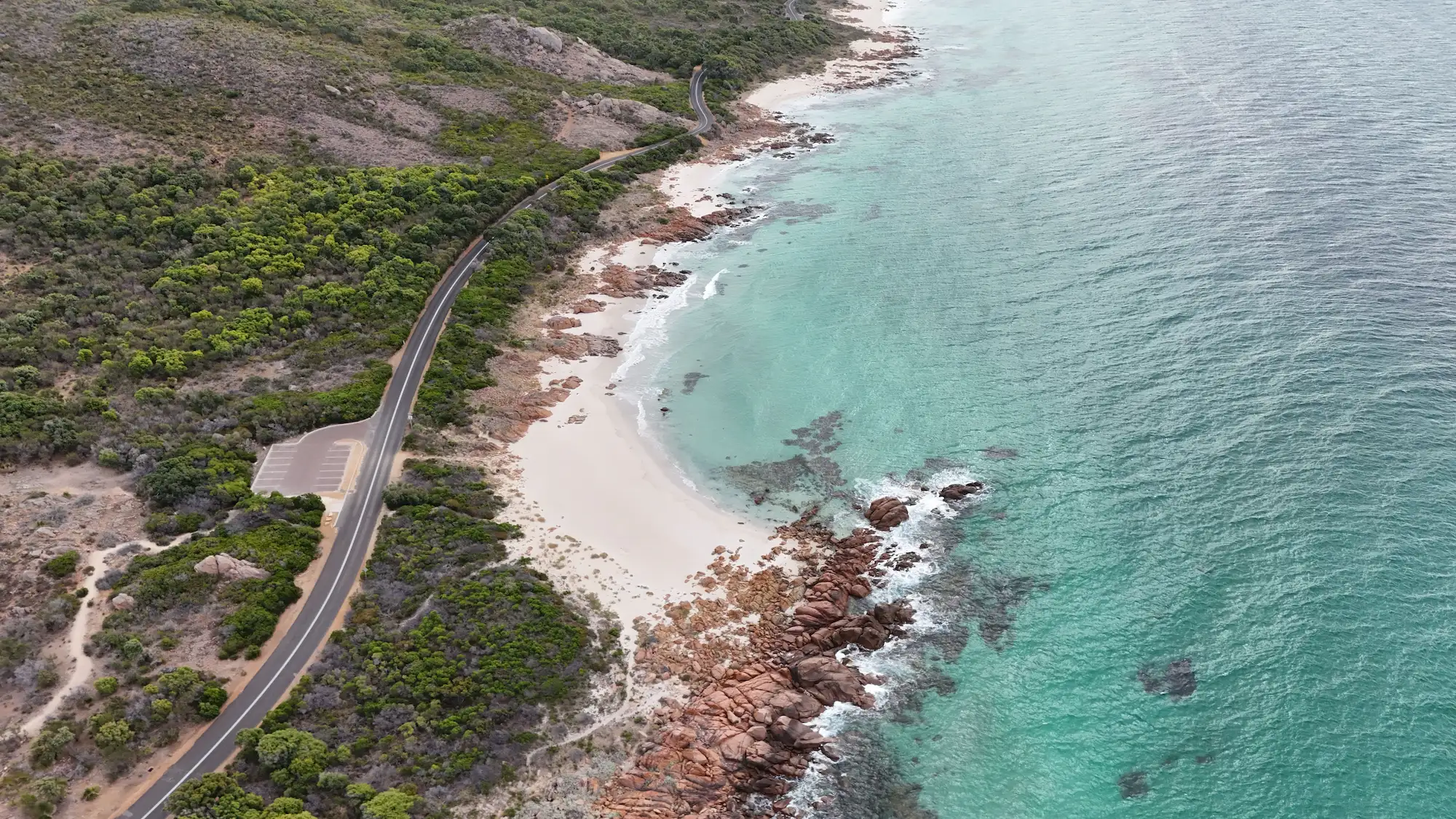 A winding coastal road next to a rocky white sandy beach with clear turquoise water.