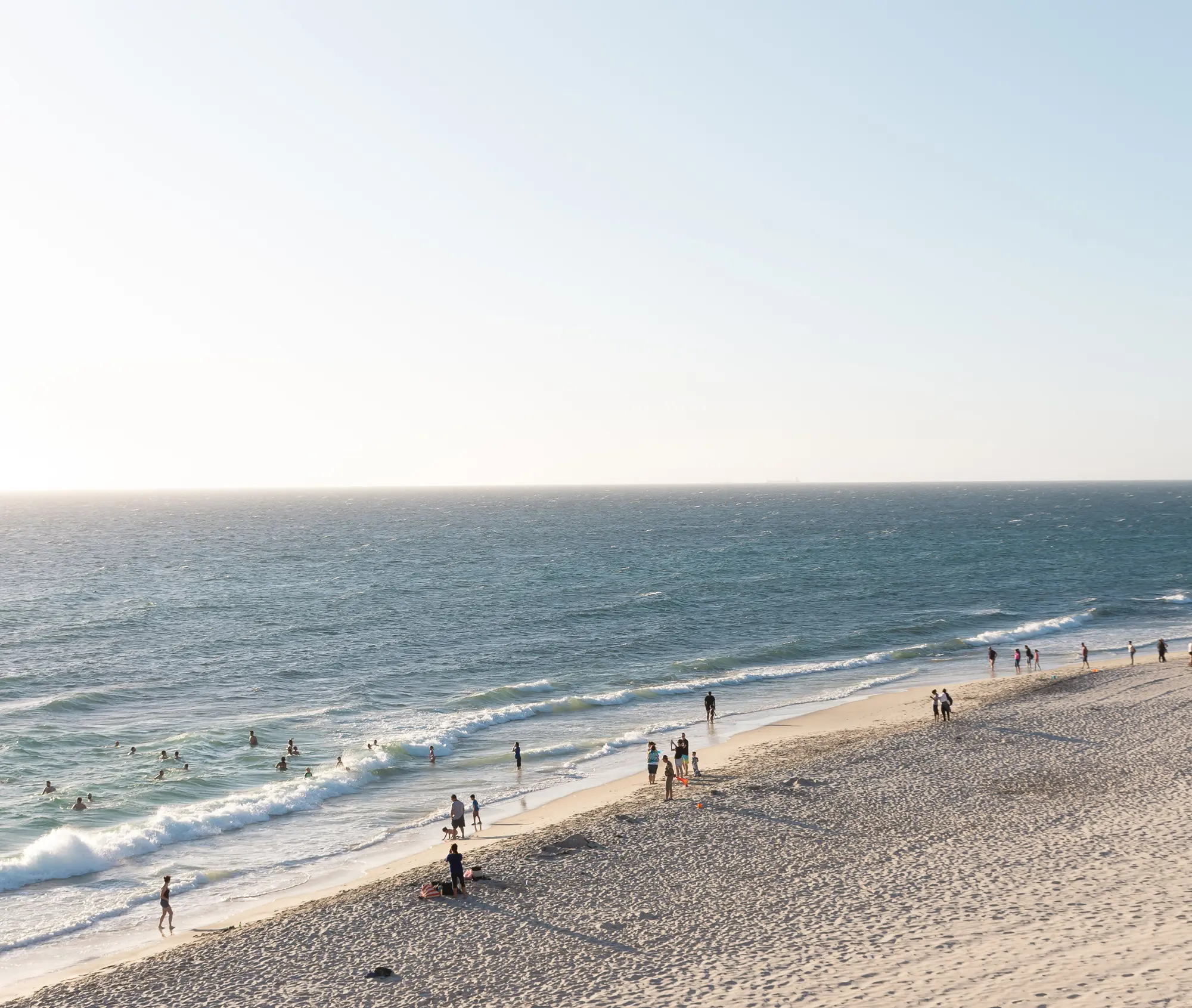 People swimming and walking along a wide sandy beach in Perth with calm waves and clear sky.