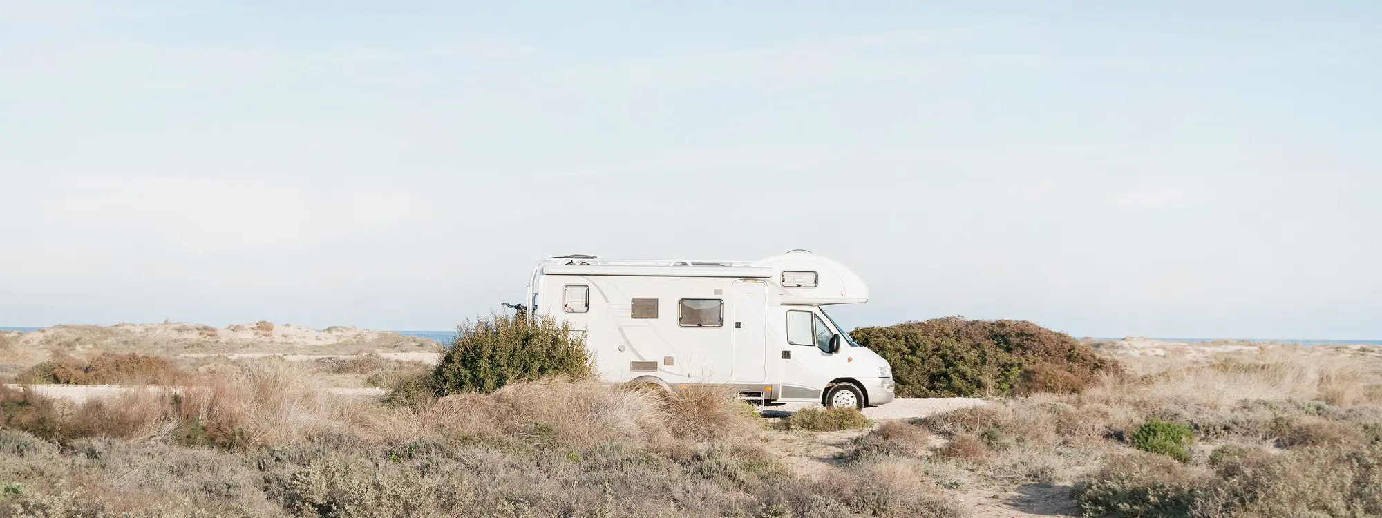 White camper van parked on a sandy path surrounded by dry grass and shrubs near the coast.