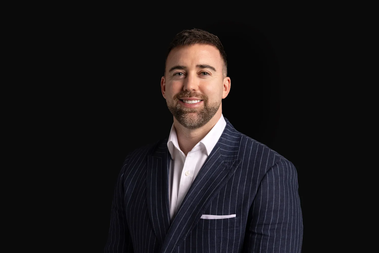 Smiling man with short hair and beard wearing a dark pinstripe suit and white shirt on black background.