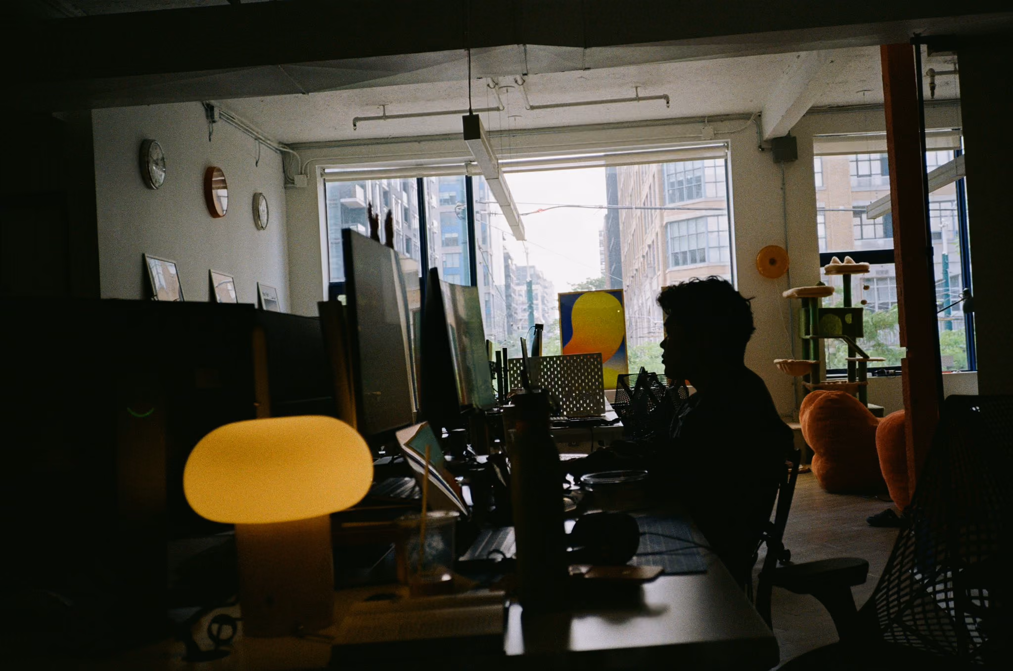 Silhouetted person working at a desk with computer monitors by a large office window in daylight.