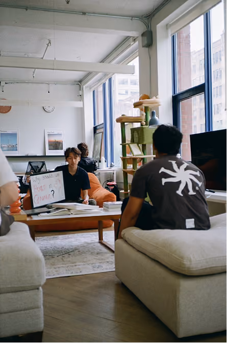 Three people in a bright living room, one holding a whiteboard that reads 'Why you? How?'