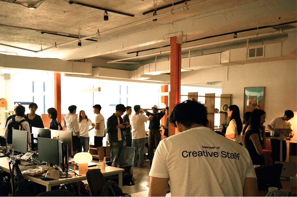 People socializing and working in a bright modern office with desks, computers, and creative staff shirt visible.