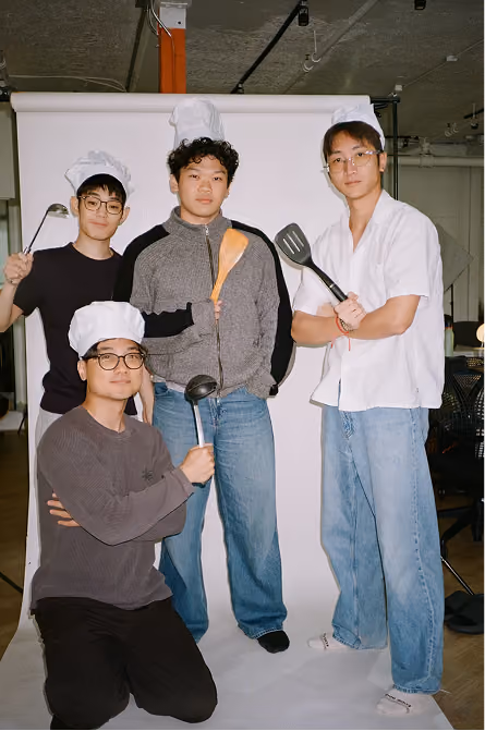 Four men wearing chef hats posing with kitchen utensils in front of a white backdrop in an indoor setting.