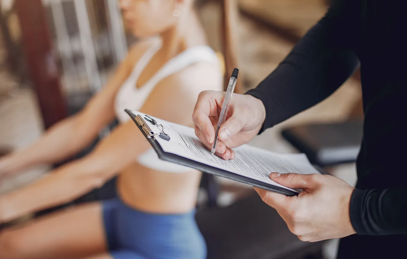 A person in a black long-sleeve shirt holding a clipboard and pen, writing on a form, with a blurred woman in workout attire sitting in the background.