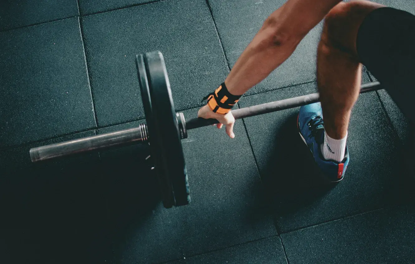 Close-up of a person in gym shoes gripping a barbell with weight plates on a rubber gym floor.