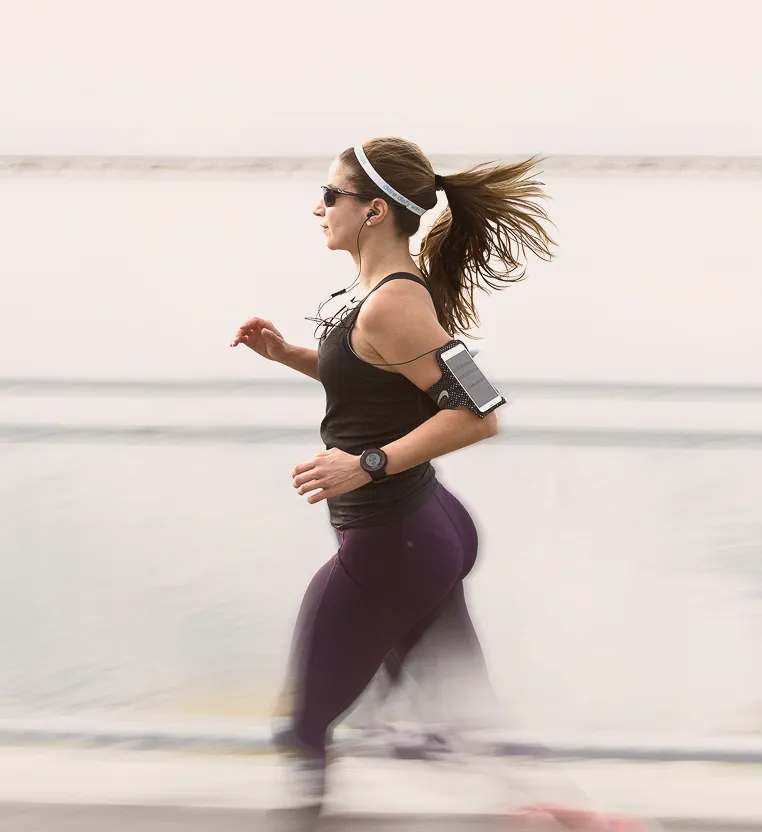 Woman with a ponytail wearing sunglasses and earbuds, running outdoors along a waterfront, with motion blur on her legs, wearing a black tank top, purple leggings, a wristwatch, and a smartphone armband.