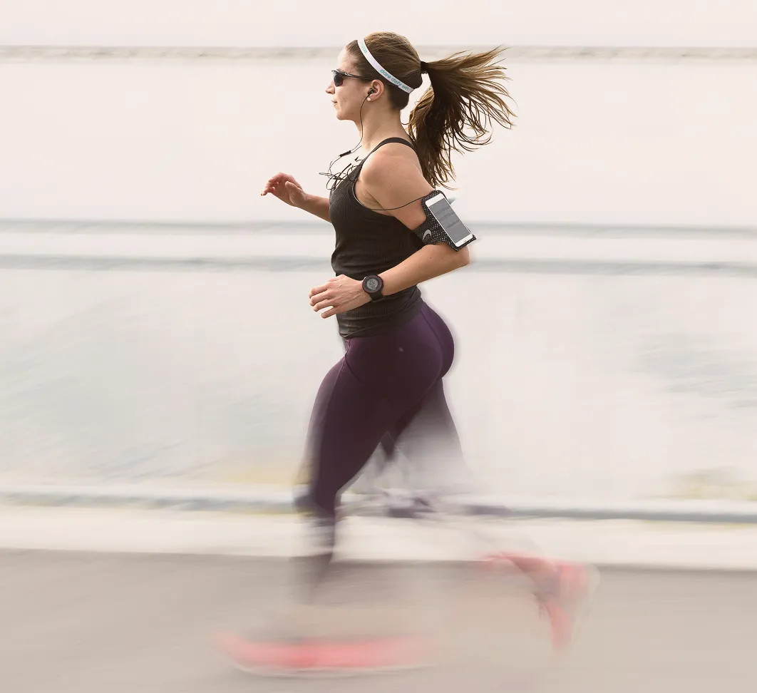 Woman running outdoors near water wearing sunglasses, wireless earphones, a headband, black tank top, and purple leggings with a smartphone in an armband and a fitness watch.