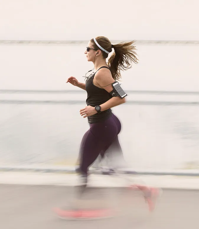 Profile of a woman jogging outdoors near water, wearing sunglasses, a black tank top, purple leggings, a fitness watch, and headphones connected to a smartphone strapped to her arm.