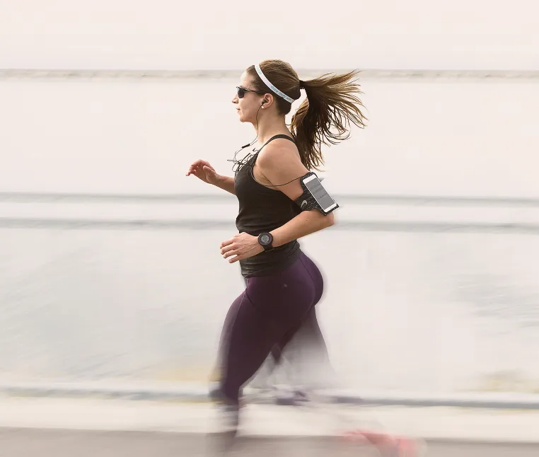 Woman jogging outdoors near water wearing sunglasses, a headband, earphones connected to a phone on her arm, and a fitness watch.