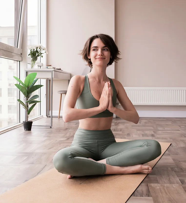 Young woman in green sportswear sitting cross-legged on a yoga mat in a bright room with large windows and plants, holding her hands in a prayer position and smiling slightly.