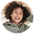 Smiling young woman with curly hair wearing a green jacket and white shirt, posing with hands behind her head against a plain white background.