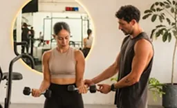A female client in a beige sports top and black leggings lifts a dumbbell while a male trainer in a dark sleeveless shirt guides her form in a gym setting with a round mirror and plants.