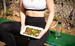 Person wearing black leggings and a white sports bra sitting outdoors on a green bench holding a white tray with a meal consisting of pieces of meat and broccoli, with a bottle of water on the bench beside them.
