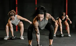 Three women in athletic wear performing a gym exercise where they bend down and extend one arm toward the floor, focusing on strength and balance in an indoor fitness setting.