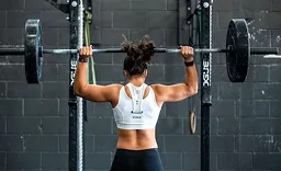 Woman with hair tied up lifting a loaded barbell overhead in a gym with a black brick wall background.