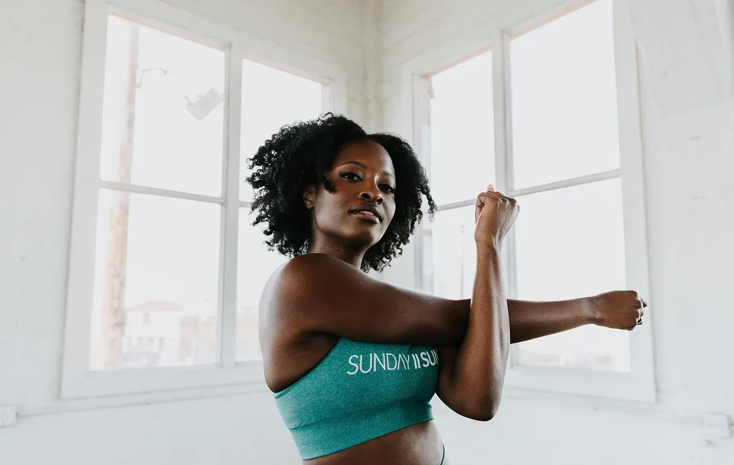 Woman in a teal sports bra stretching her arms in a bright room with large windows.