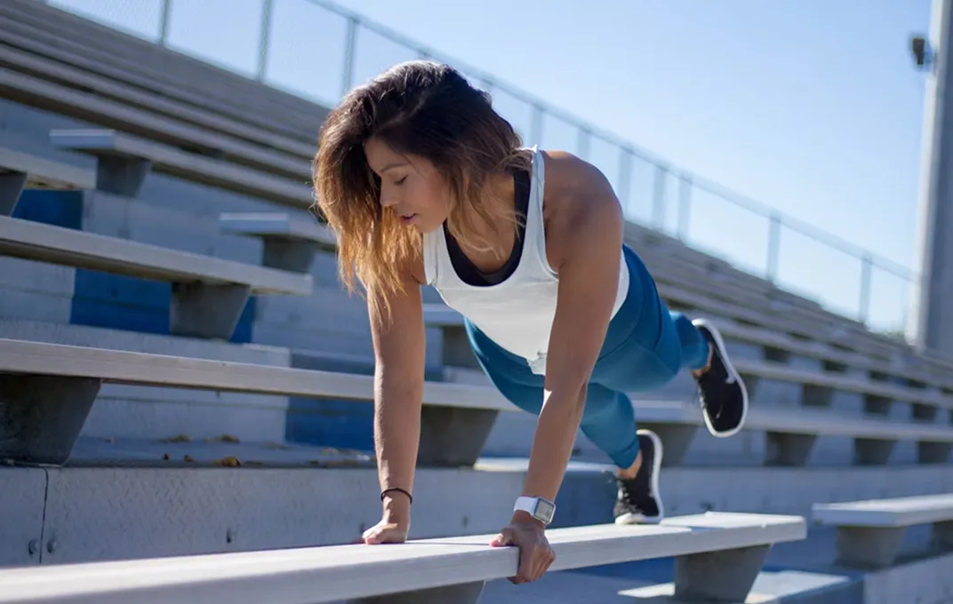 Woman in athletic wear doing elevated plank exercise with one leg raised on outdoor bleachers under clear sky.