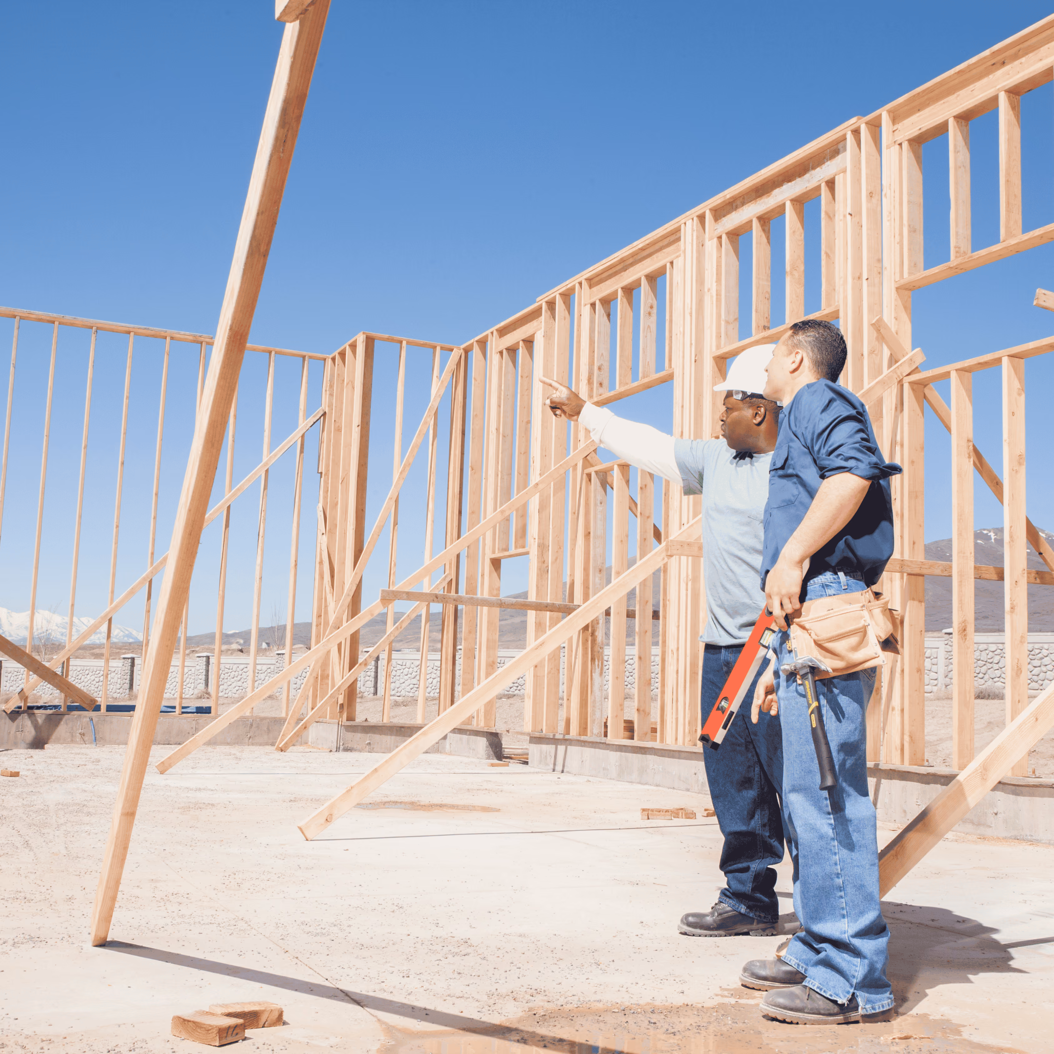Two construction workers discuss framing of a new house under a clear blue sky.