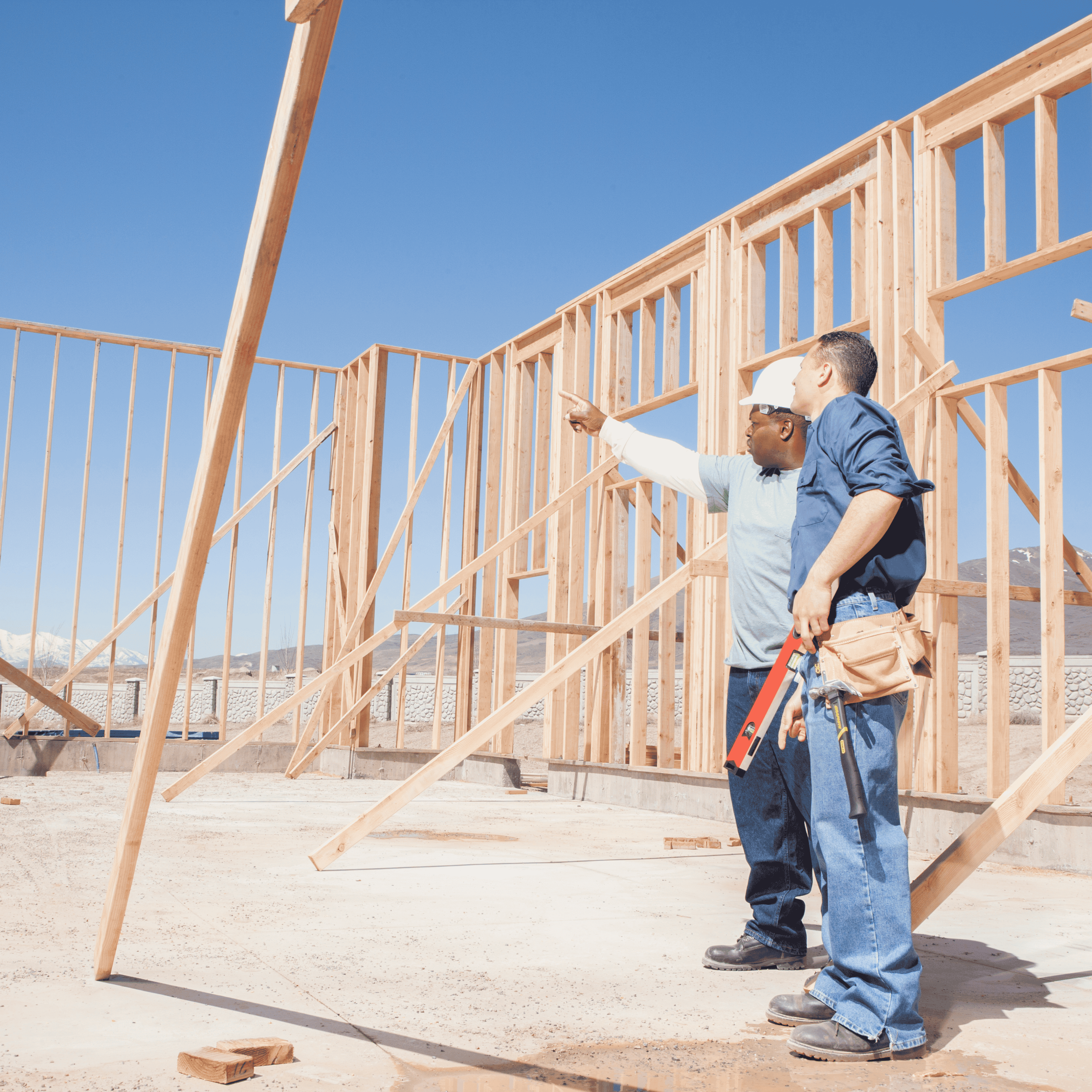 Two construction workers discuss framing of a new house under a clear blue sky.