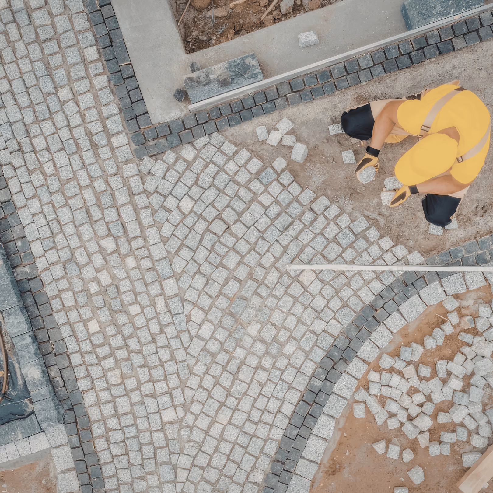 Worker in yellow outfit arranging small square stones on the ground for custom paving or hardscaping.