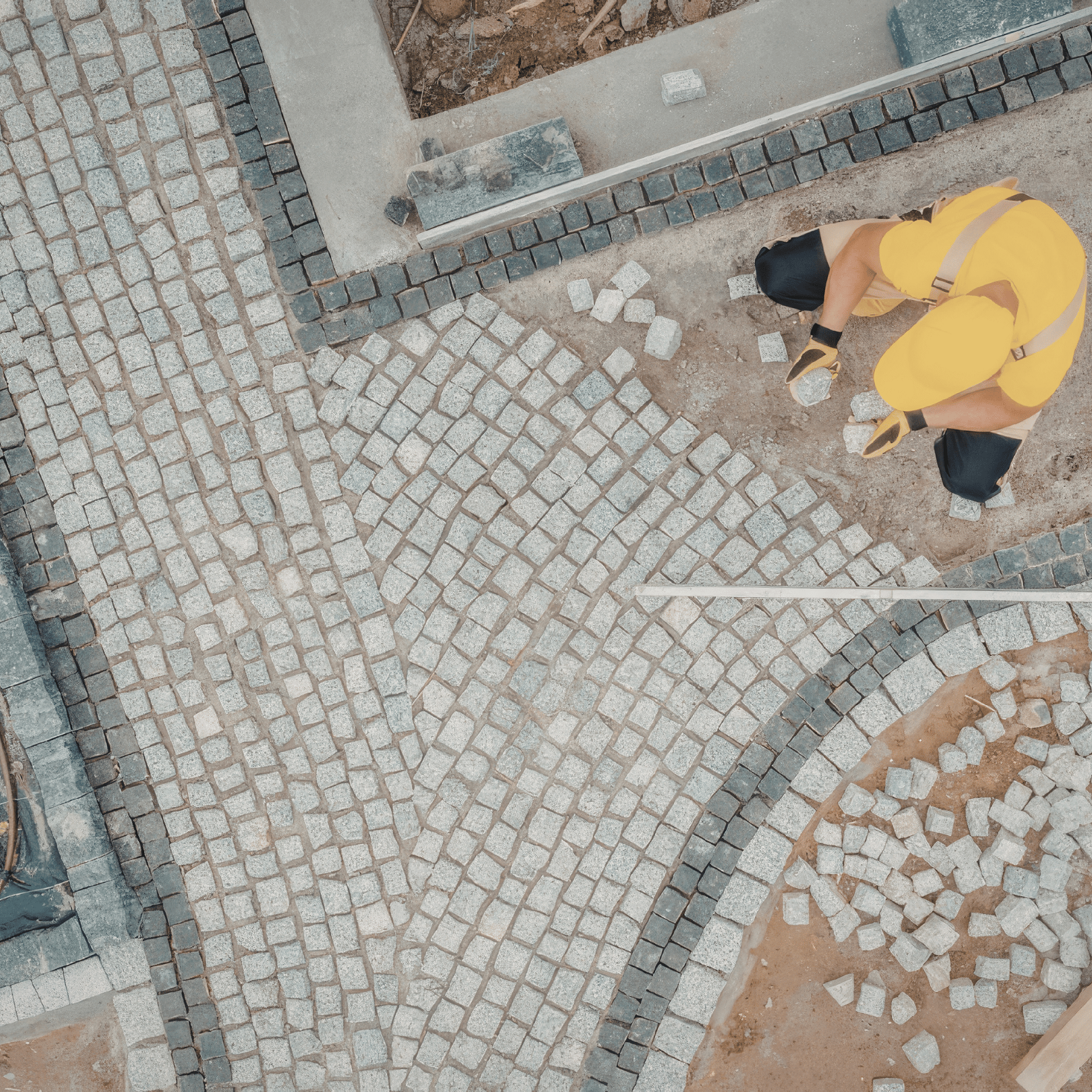 Worker in yellow outfit arranging small square stones on the ground for custom paving or hardscaping.