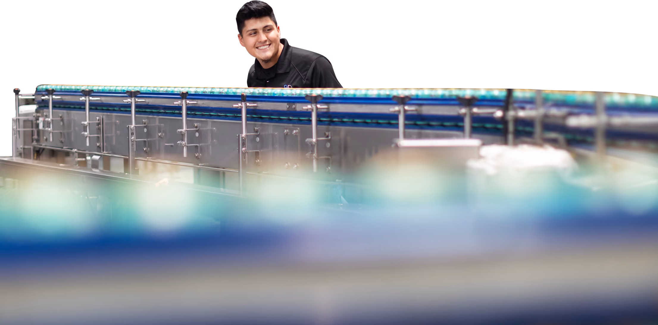 Man in black shirt smiling behind a long industrial conveyor belt with many blue capped bottles.