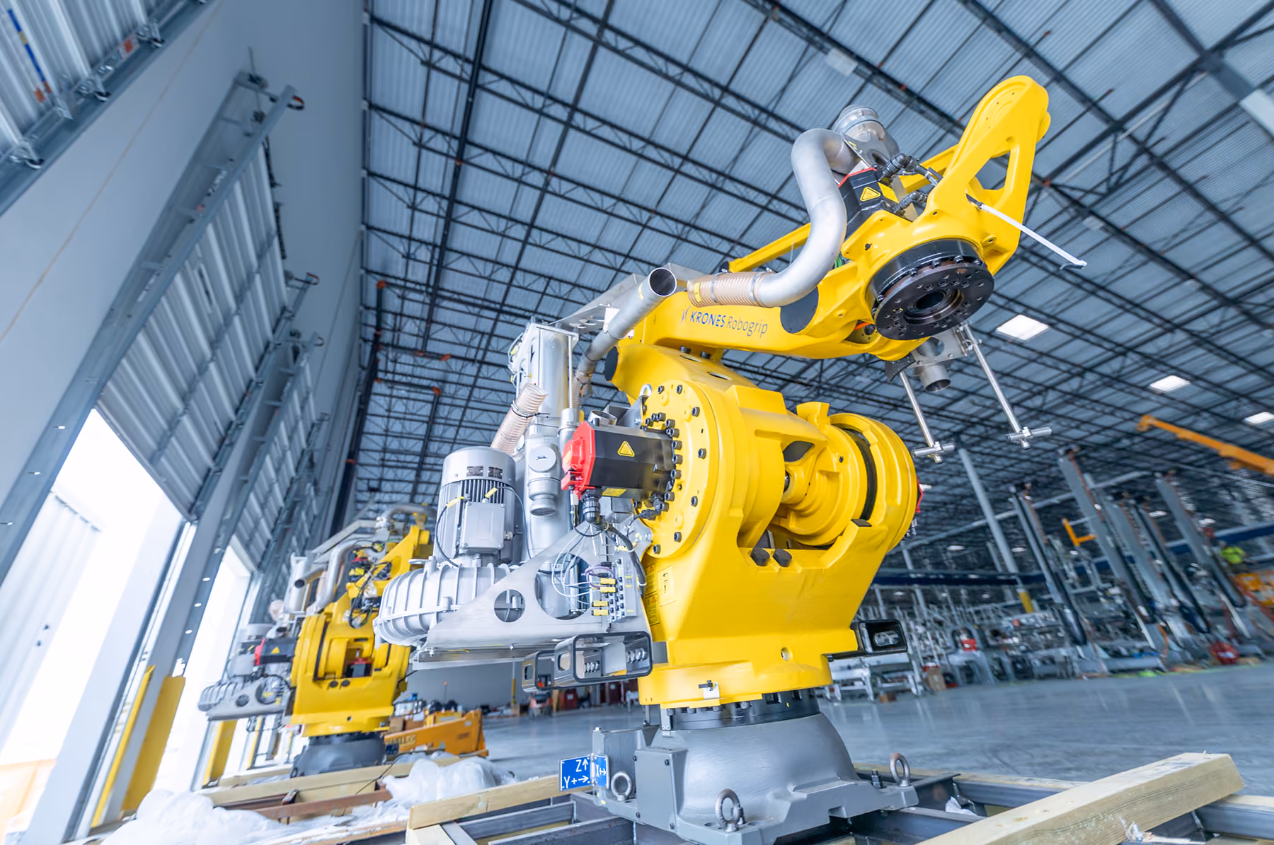 Large yellow industrial robots inside a spacious factory or warehouse with metal ceiling beams.