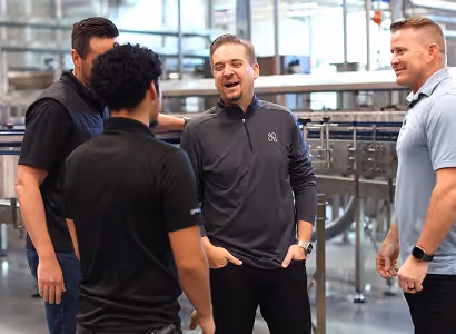 Four men standing and conversing in a bright industrial or warehouse setting.