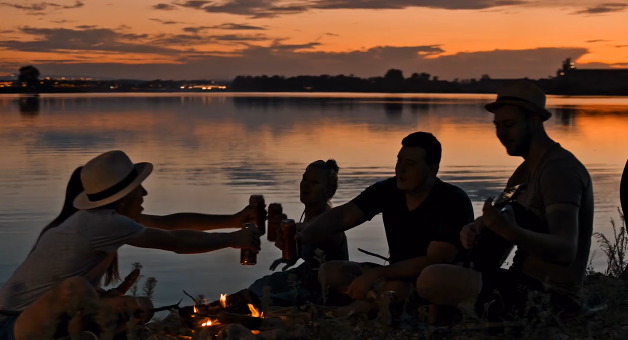 Group of friends around campfire by a lake at sunset, toasting drinks and playing guitar.