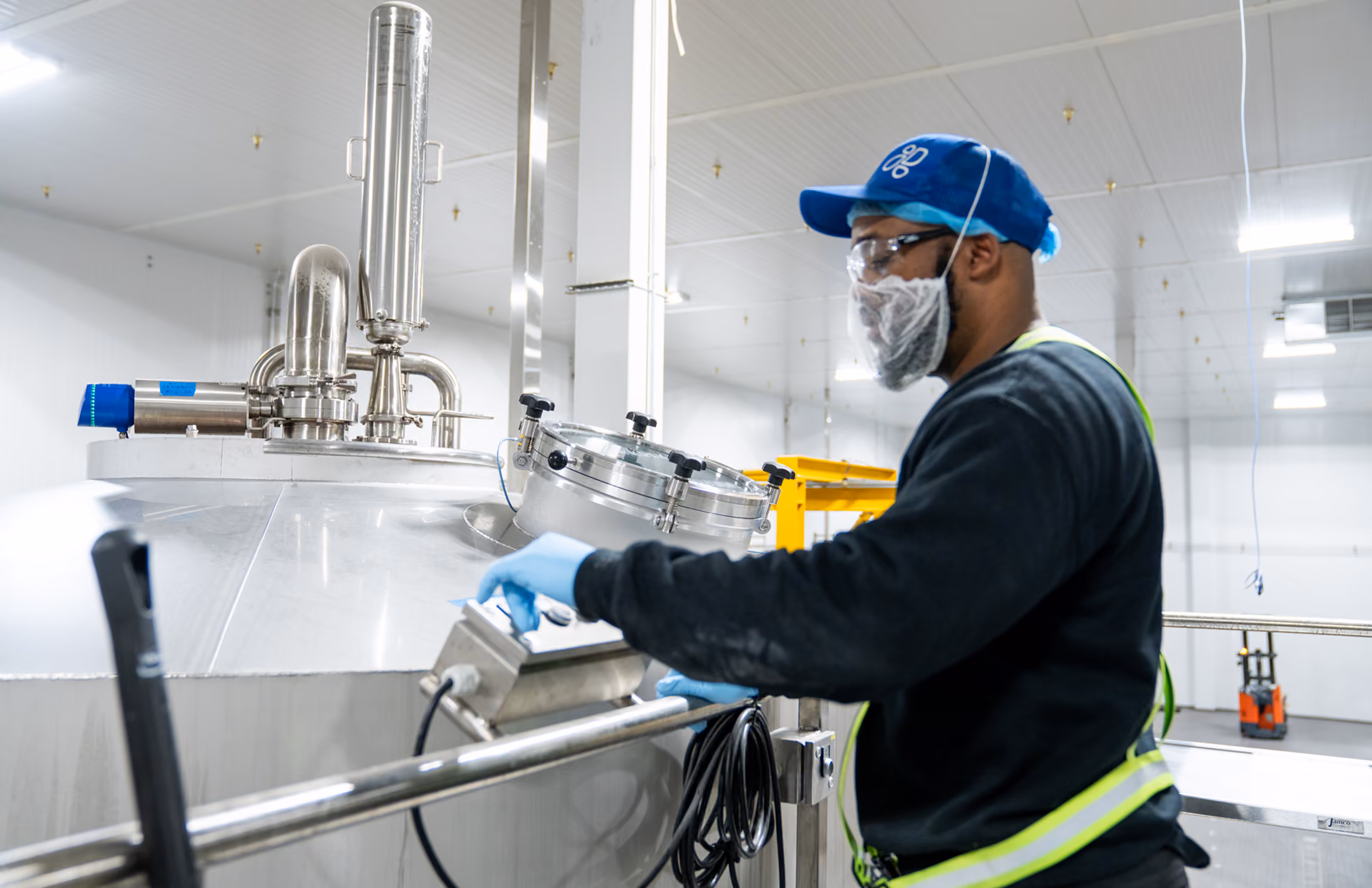 Worker in safety gear operating control panel on large stainless steel industrial tank indoors.