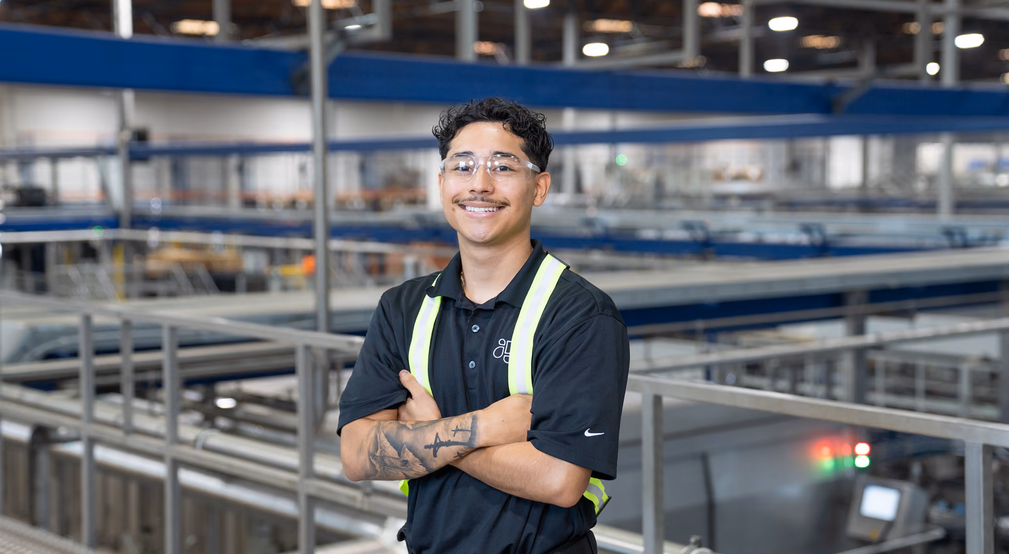 Smiling worker with safety glasses and reflective vest standing in large industrial warehouse.